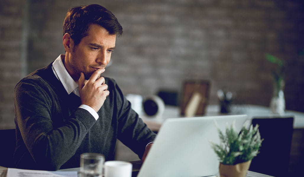 A man sits at a desk, resting his chin on his hand, while working on a laptop.