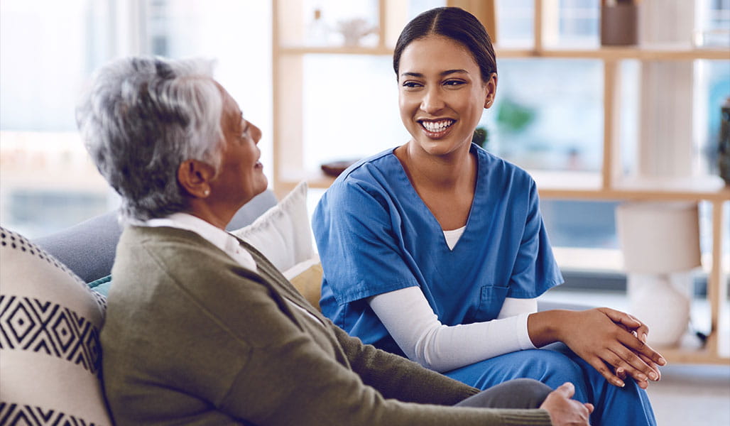 A healthcare professional sits on a couch with an elderly patient engaging in a lighthearted conversation.