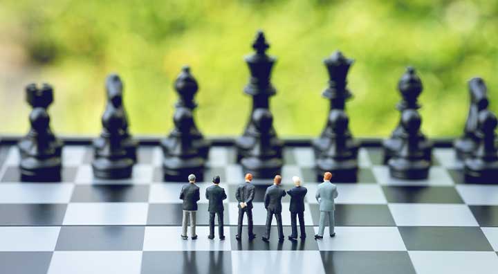 Group of miniature people businessmen standing on chessboard looking at black chess enemy in far background.