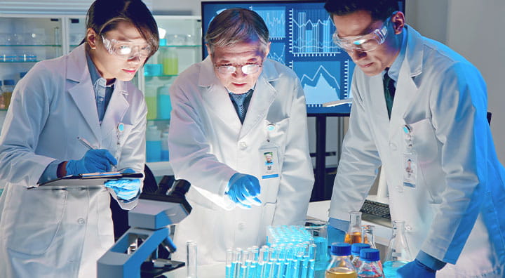 A group of scientists in lab coats studies samples in a laboratory with graphs displayed in the background.