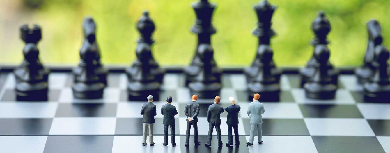 Group of miniature people businessmen standing on chessboard looking at black chess enemy in far background.