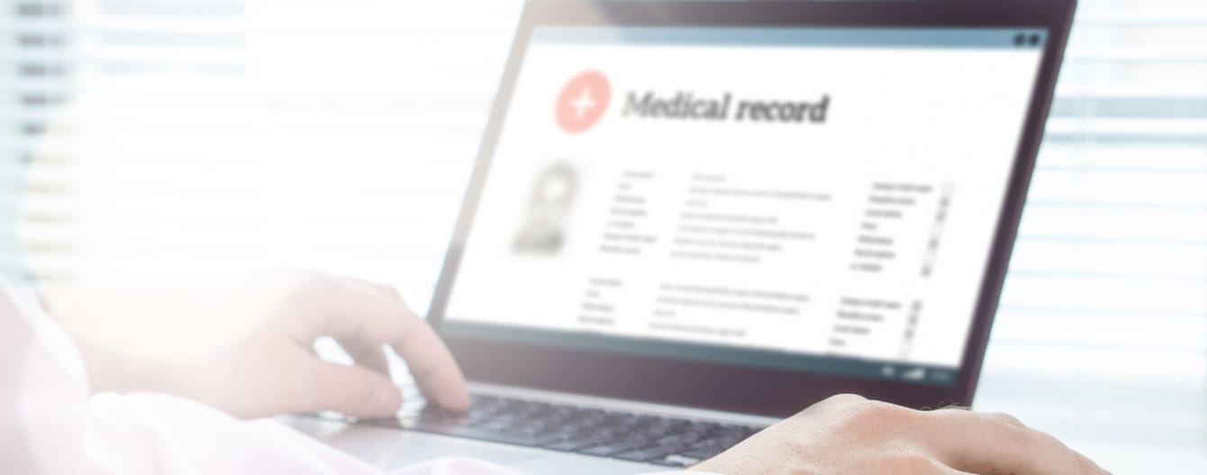 Close-up of a person in a white coat using a laptop displaying a medical record, with hands on the keyboard and mouse, in a brightly lit office.